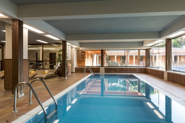 An indoor swimming pool with clear glass walls, wooden pillars, and a view of a building outside.