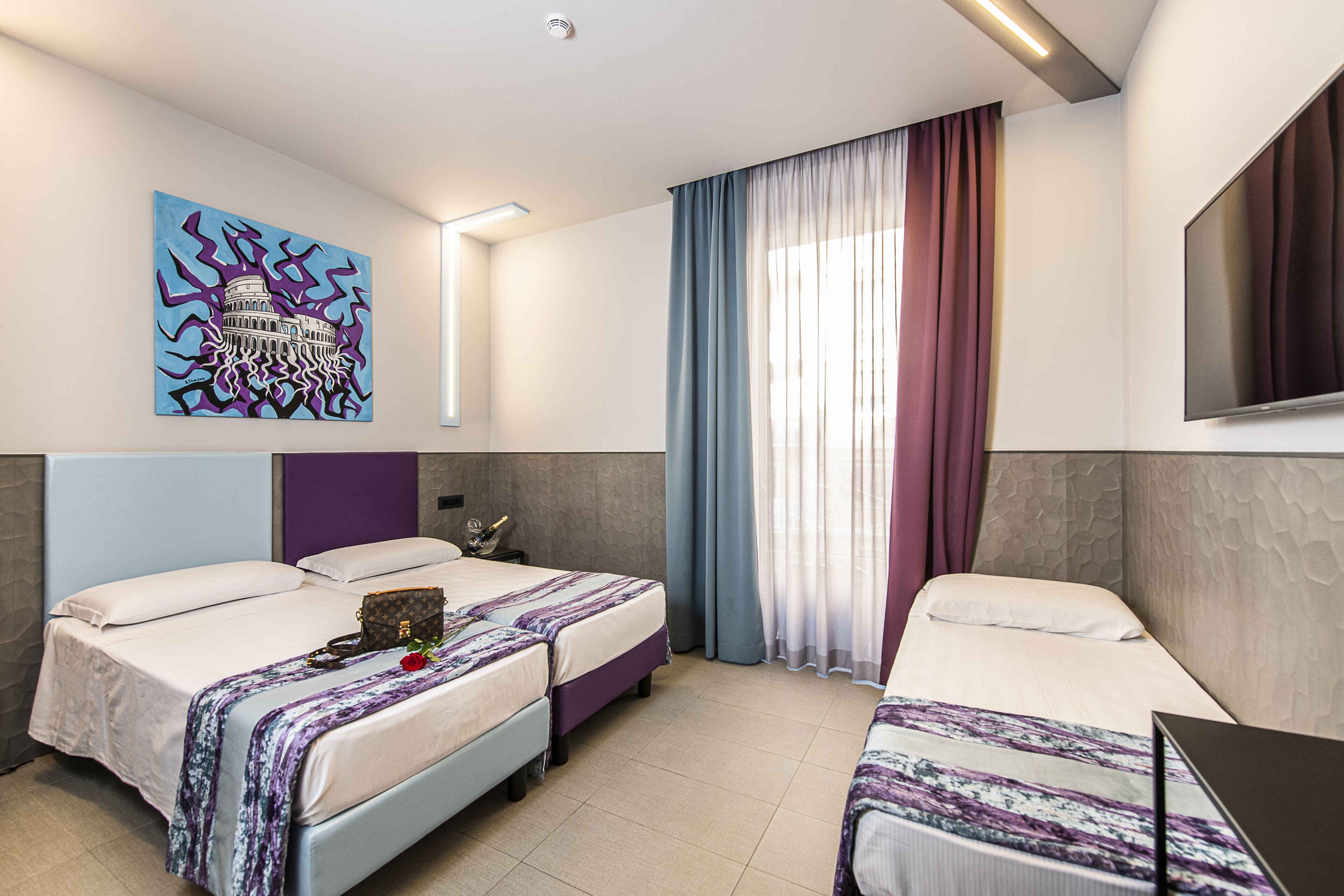 Una camera d'albergo con due letti, un quadro raffigurante un edificio, una TV e una finestra con tende.