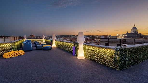 Une terrasse sur le toit, aménagée avec des sièges, un panneau d’accueil et une vue sur la ville au crépuscule.