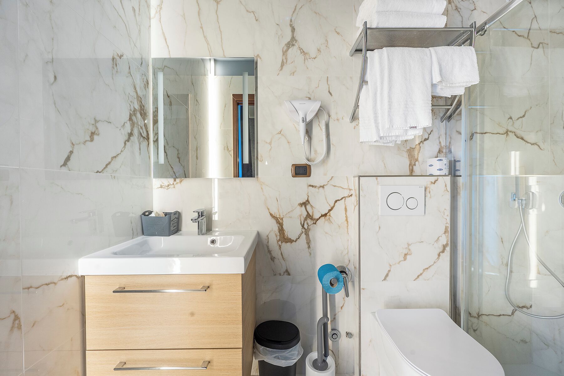 Un baño moderno con salpicadero de mármol, un lavabo blanco, un espejo, un perchero, una zona de ducha y un inodoro.