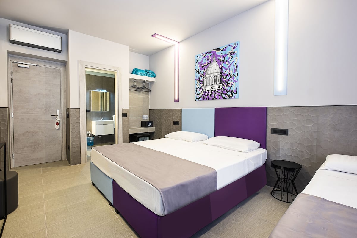 A modern hotel room with a large bed, a small table, a wardrobe, and a bathroom visible through an open door.
