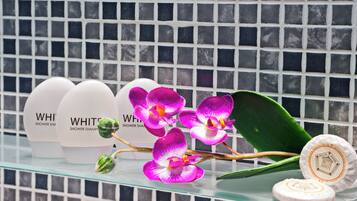 Uno scaffale in bagno con flaconi di shampoo da doccia bianchi, orchidee viola e una foglia verde.