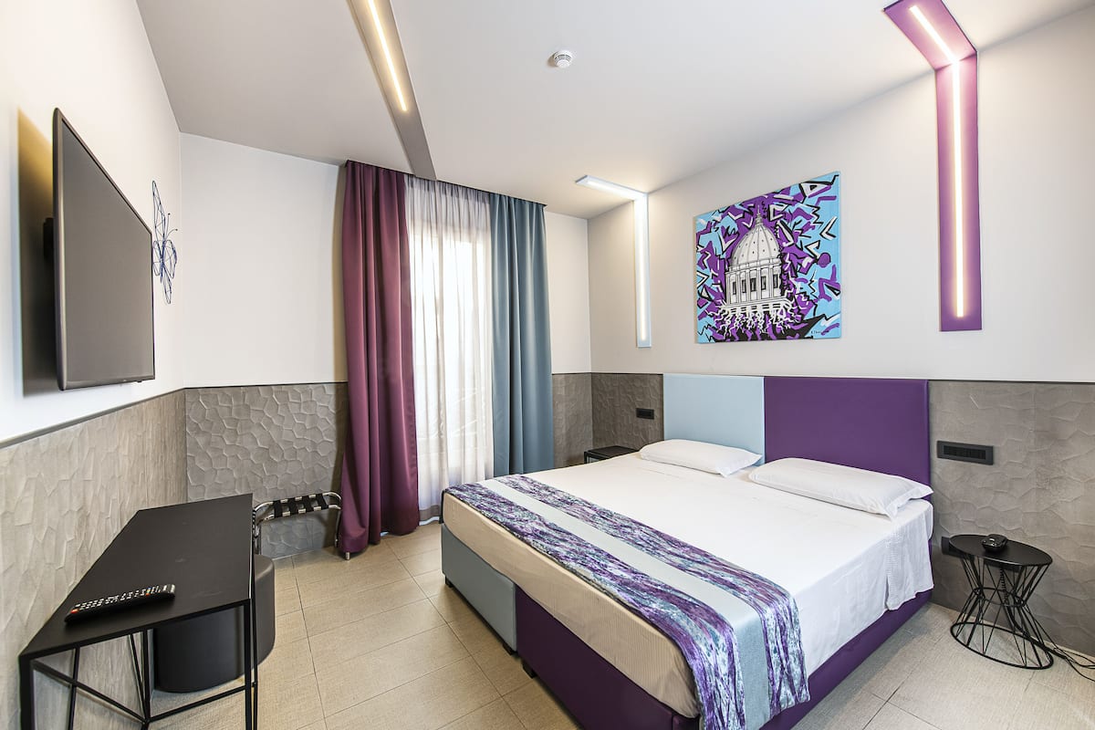 A modern hotel room with a large bed, a desk with a computer, a TV, and a painting on the wall.