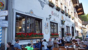 Uma estalagem bávara tradicional com mesas ao ar livre, janelas amplas com caixas de flores e uma placa indicando hotel e restaurante.
