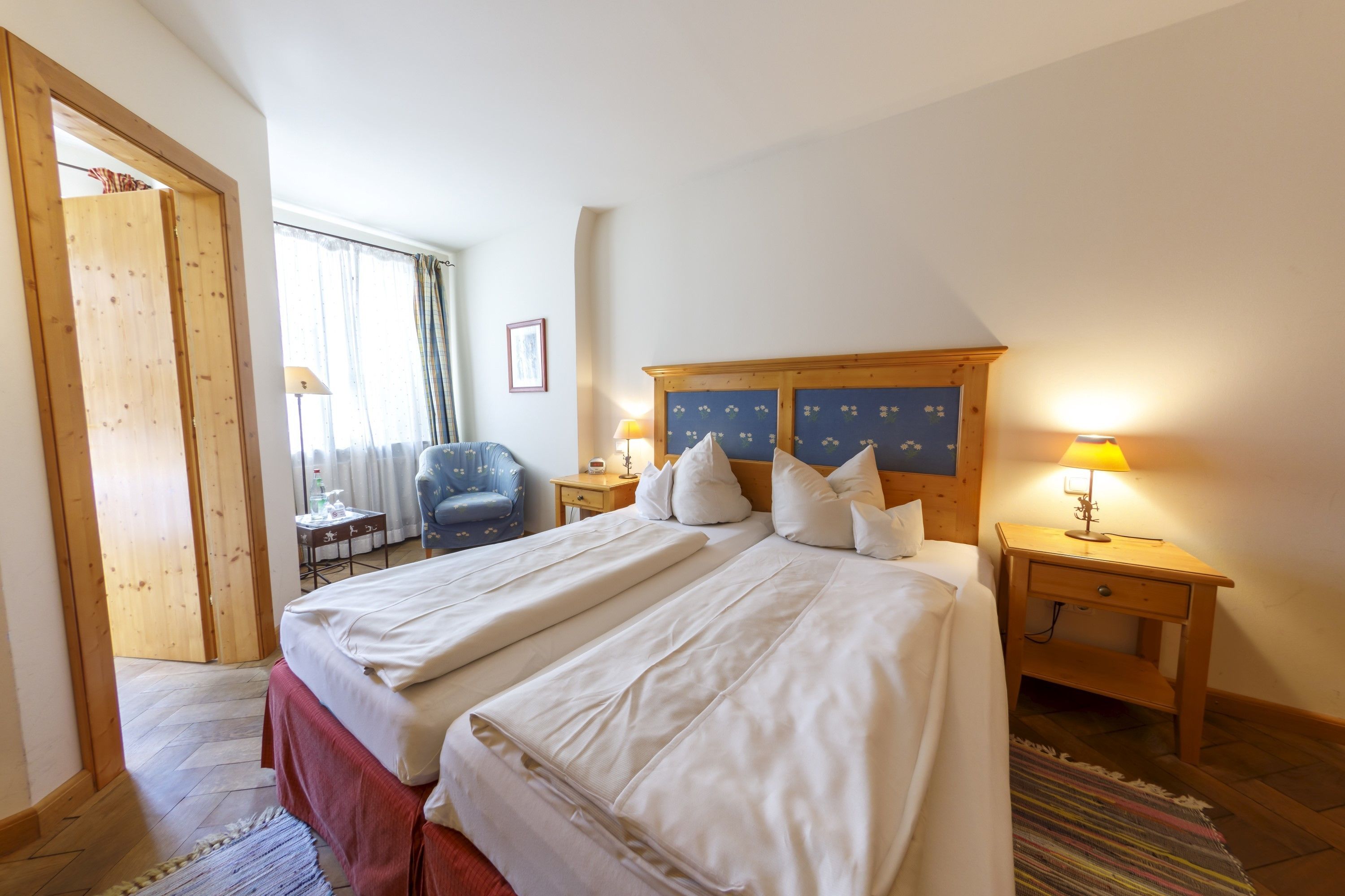 Une chambre d’hôtel avec deux lits, une tête de lit en bois, des lampes de chevet, une chaise, une petite table et une porte donnant sur une autre pièce.