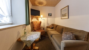 Um quarto aconchegante com teto de madeira, um sofá, uma poltrona, uma mesa pequena com papéis, um abajur e uma televisão na parede.
