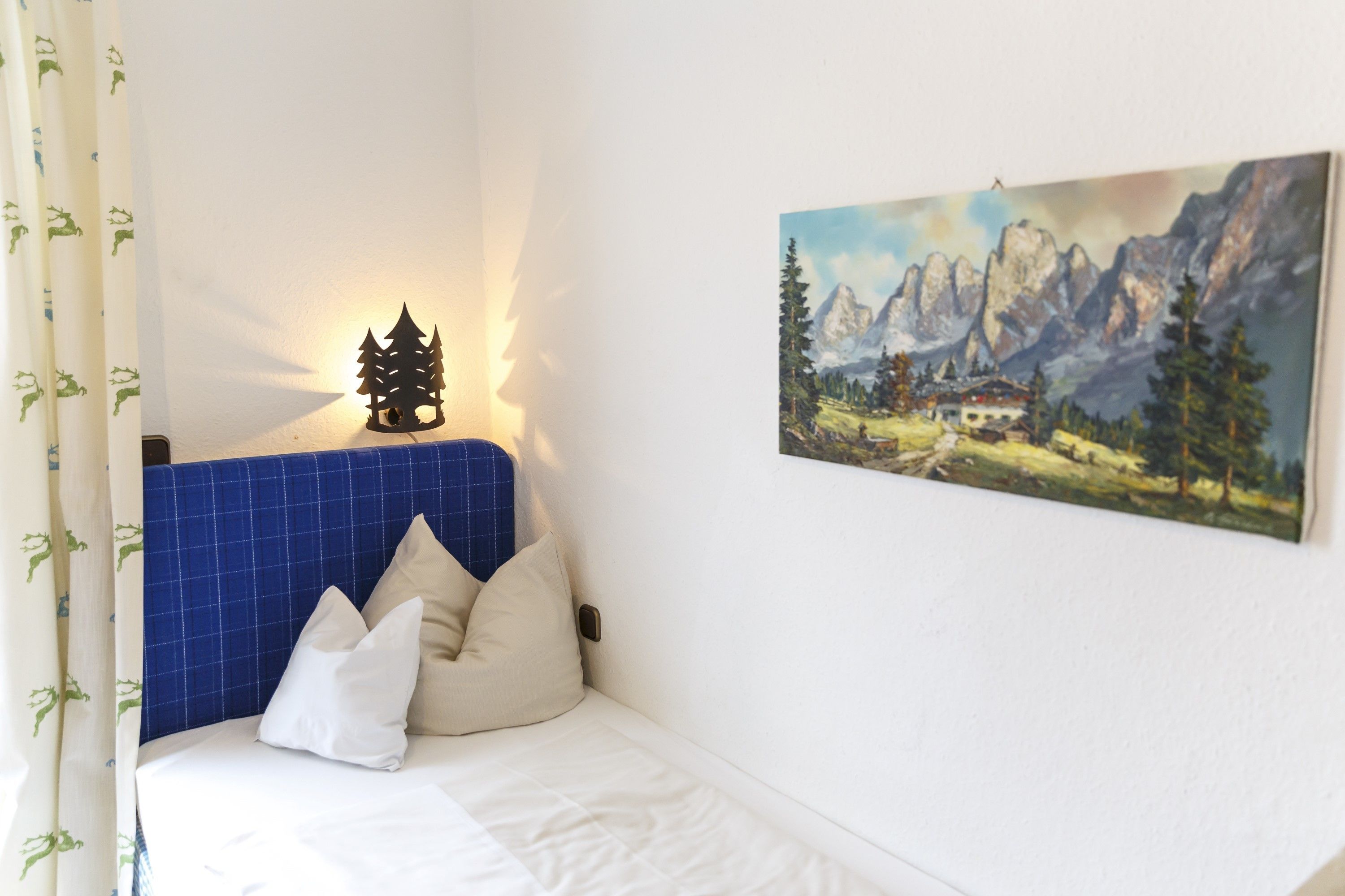 A bedroom with a bed, white bedding, a blue headboard, a painting of mountains, and a wall-mounted light fixture.