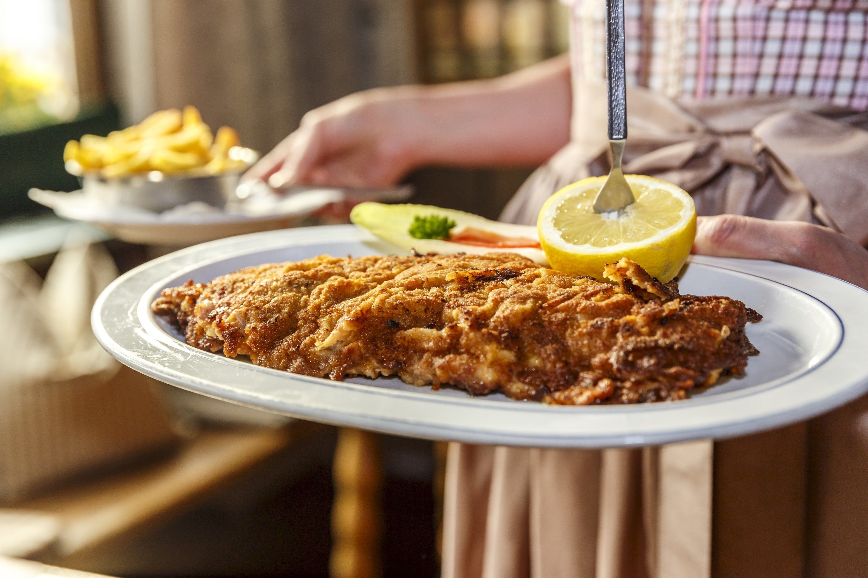 Una persona sirve un pescado frito grande y crujiente en un plato con limón y verduras.