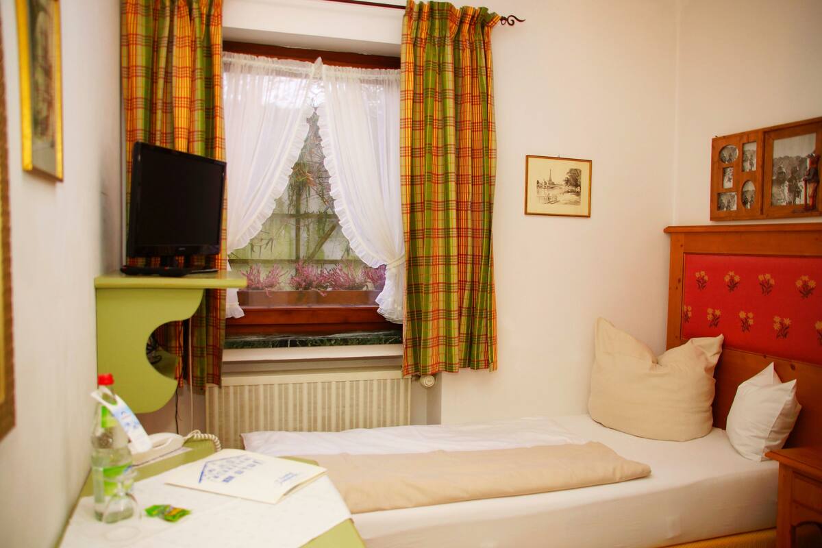 Een slaapkamer met een bed, een nachtkastje, een televisie op een standaard, een raam met gordijnen en een muur met ingekaderde foto’s.