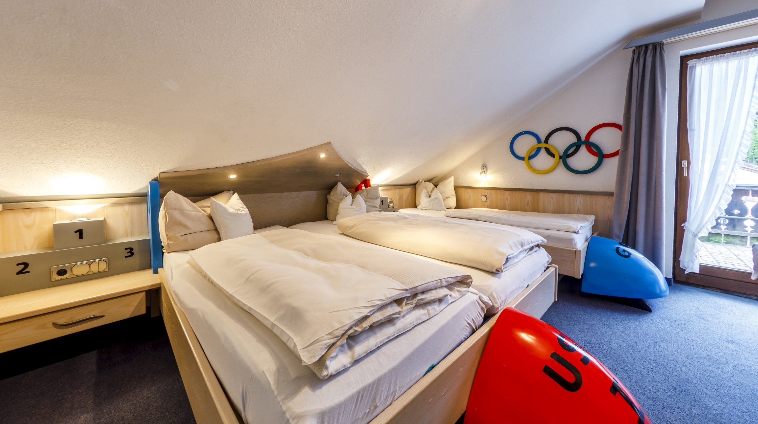 A hotel room with two beds, a desk, and a chair. There are Olympic rings on the wall and a red suitcase on the floor.