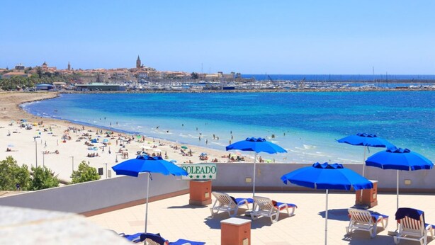 A beach with sun loungers and umbrellas, a clear blue sea, and a distant view of buildings. - Hotel Soleado (Alghero)