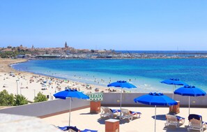A beach with sun loungers and umbrellas, a clear blue sea, and a distant view of buildings. - Hotel Soleado (Alghero)