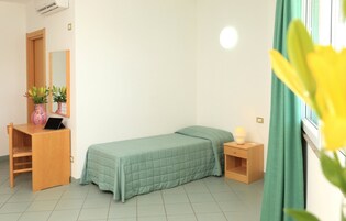 Una habitación pequeña y ordenada con una cama individual, un escritorio con una computadora portátil, un espejo y una mesita de noche con una lámpara.