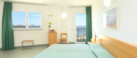 A hotel room with a bed, a desk, a chair, a small table with a vase of flowers, a picture on the wall, and a balcony with a view of the sea.