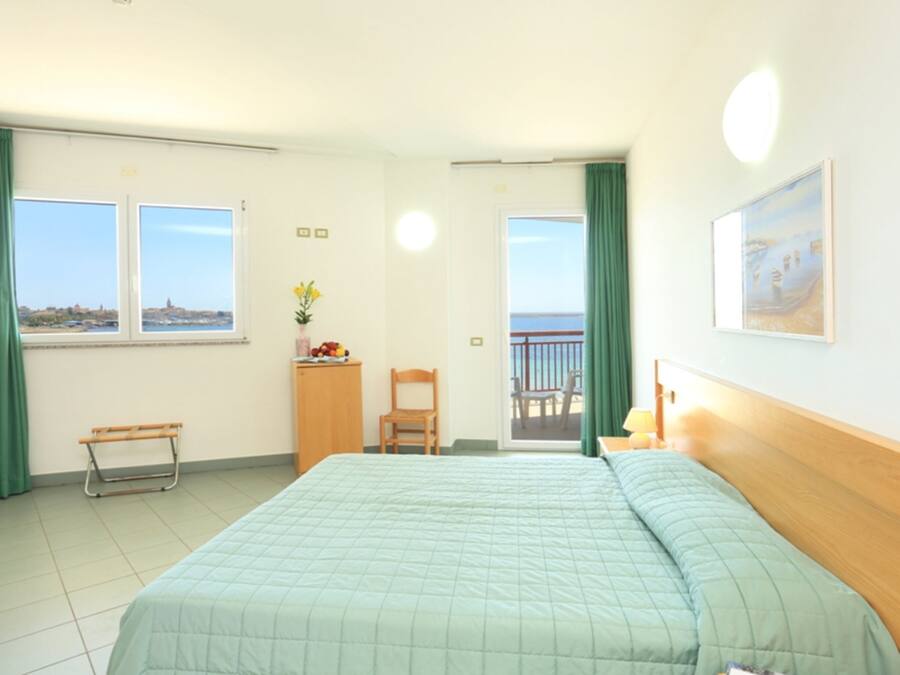 Habitación de hotel con cama, escritorio, silla, pequeña mesa con un florero, un cuadro en la pared y balcón con vista al mar.