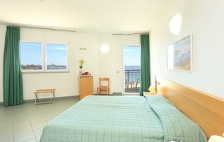 A hotel room with a bed, a desk, a chair, a small table with a vase of flowers, a picture on the wall, and a balcony with a view of the sea.