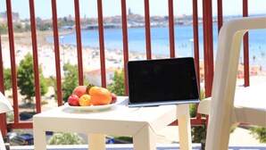 A laptop on a white table with a plate of fruit and a view of a beach and sea in the background. - Hotel Soleado (Alghero)