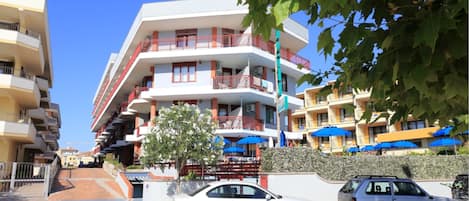 A multi-story residential building with balconies, a parking area, and blue umbrellas.