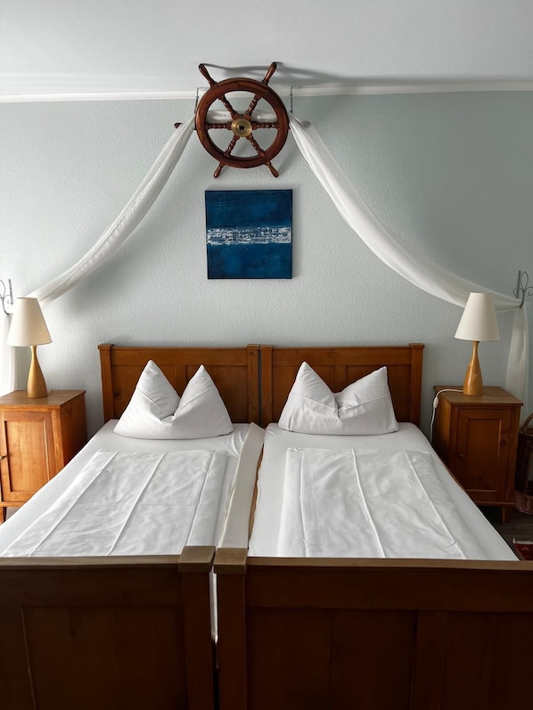Uma cama de casal com lençóis brancos, uma cabeceira de madeira e duas mesas de cabeceira com luminárias. Uma decoração de leme de navio está pendurada acima da cama, e um quadro abstrato azul na parede.