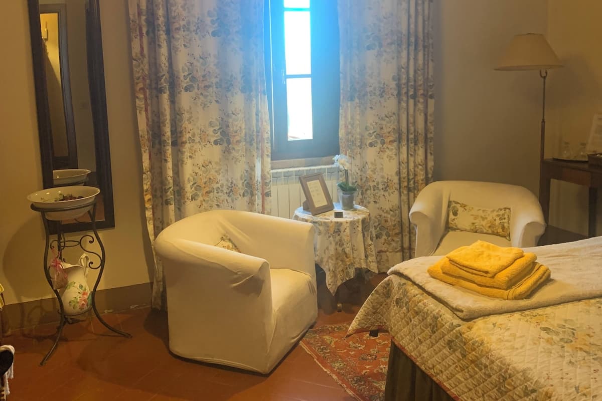 A room with a bed, two chairs, a small table, a mirror, and a window with curtains.