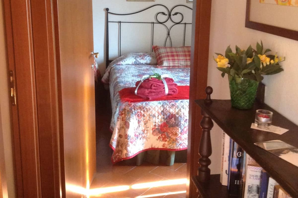 A bedroom with a bed, a bedside table with a vase of flowers, a bookshelf, and a door.