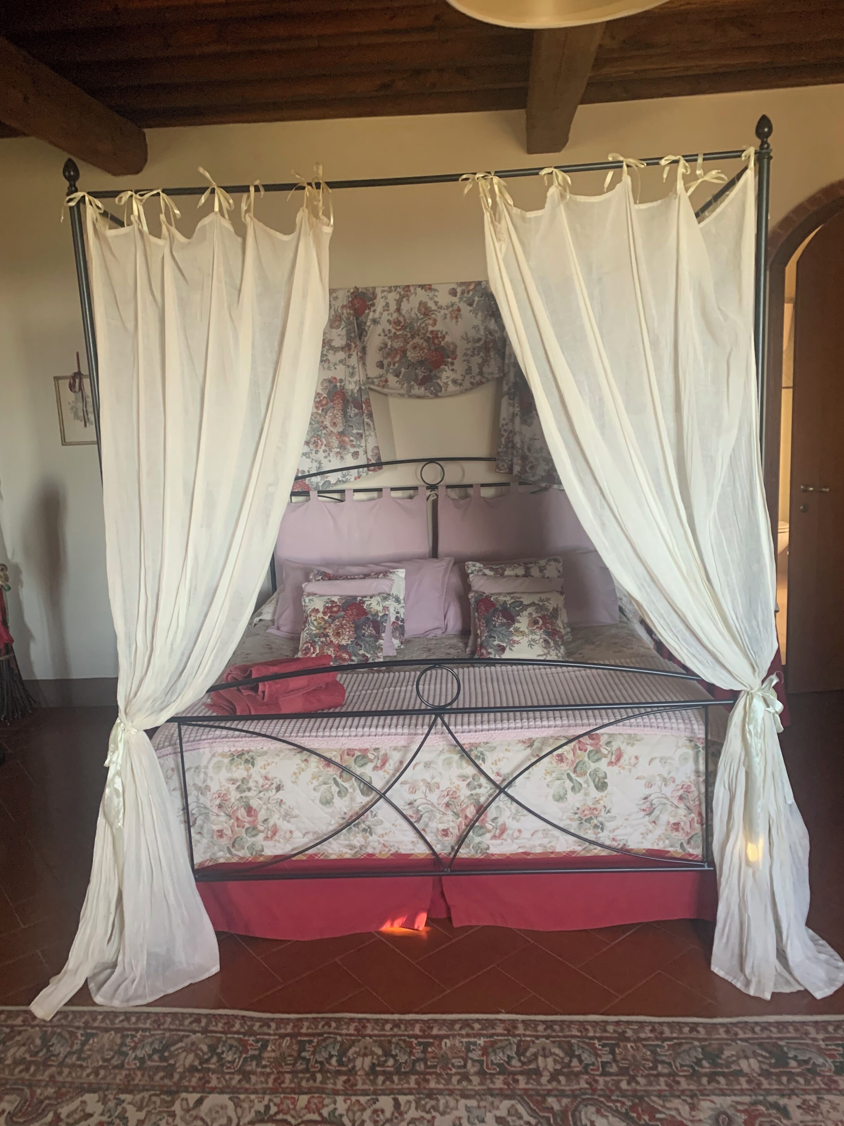 Een hemelbed met bloemenbeddengoed en een rode sprei, een kamerbreed tapijt met dessin, en een houten plafond.