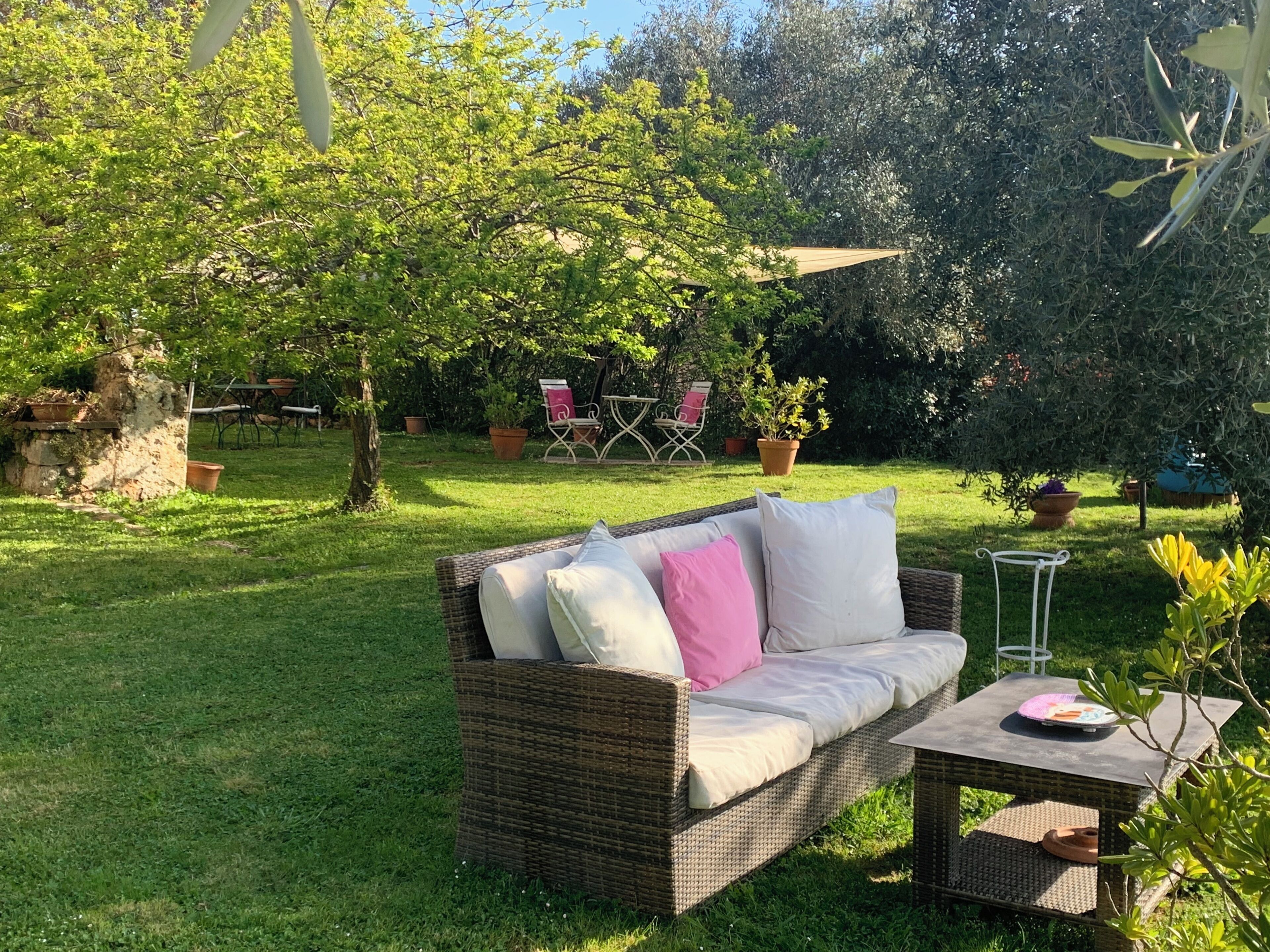 Un sofá de mimbre con cojines, una mesita y una silla en un jardín con césped, plantas en macetas y una pérgola.