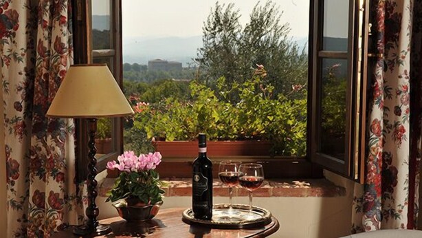 A table with a bottle of wine, two glasses, a lamp, and a potted plant by a window with floral curtains. - La Canonica di San Michele (Monteriggioni)