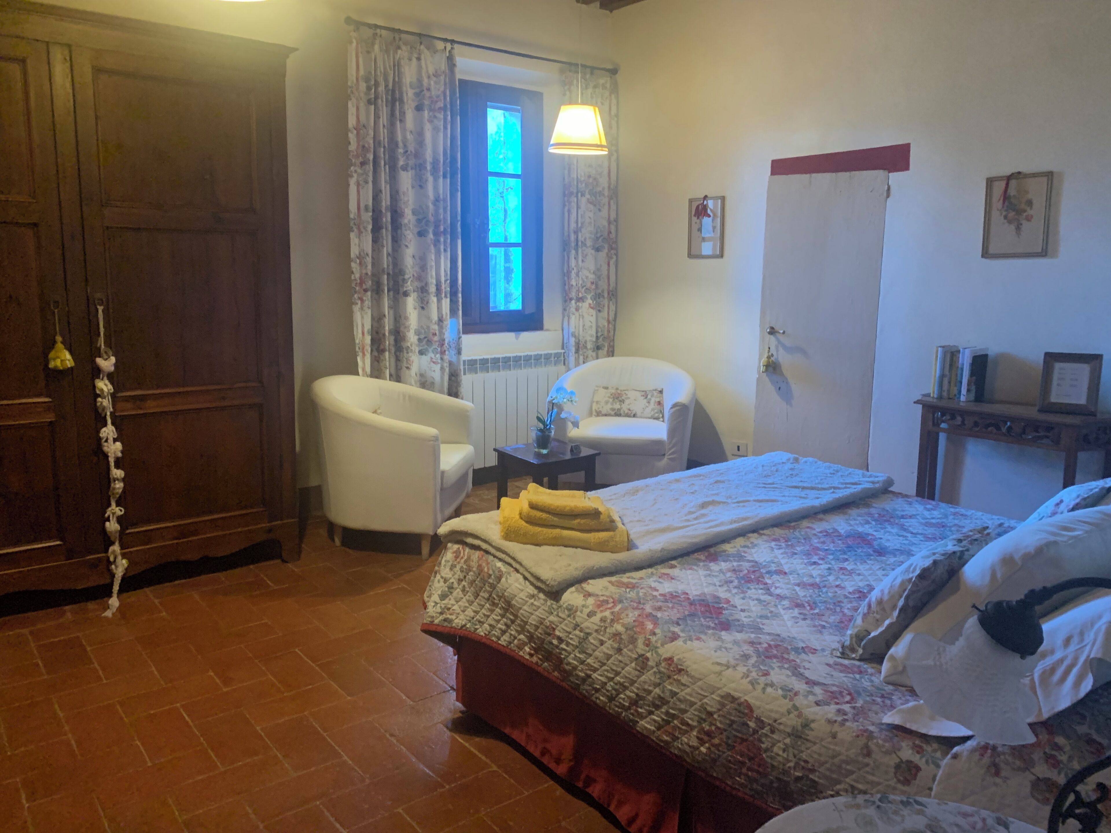 Un dormitorio con cama, dos sillas, una ventana con cortinas, una puerta de madera y una mesita con lámpara.