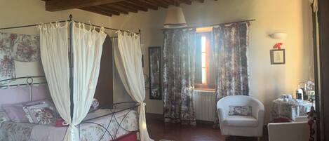 A bedroom with a four-poster bed, a sitting chair, a small table with a lamp, and a window with curtains.
