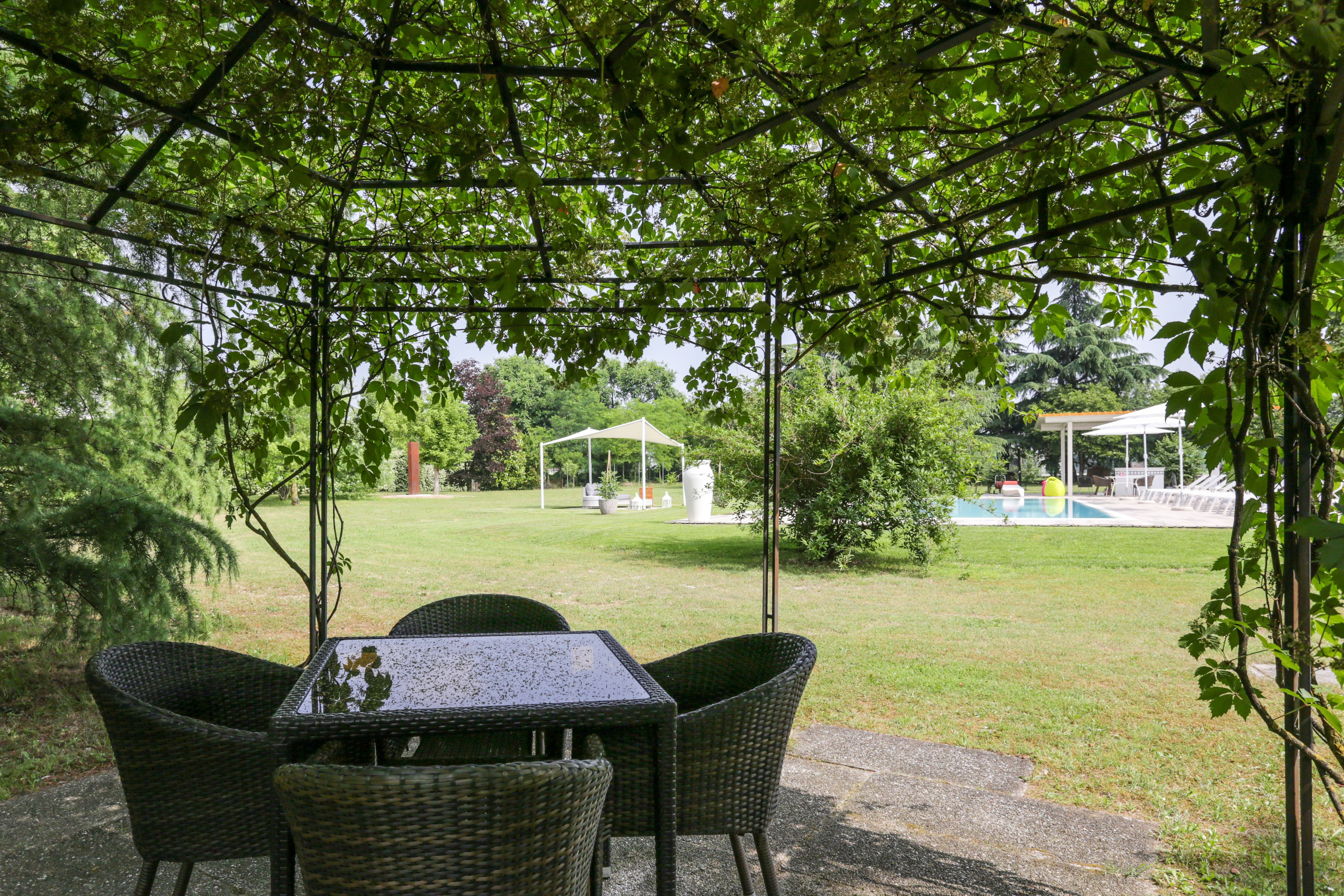 A covered outdoor seating area with wicker chairs and a table, overlooking a grassy lawn and a pool area.