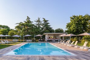 Une piscine entourée d’arbres, avec des chaises longues et des parasols, ainsi qu’un espace bar.
