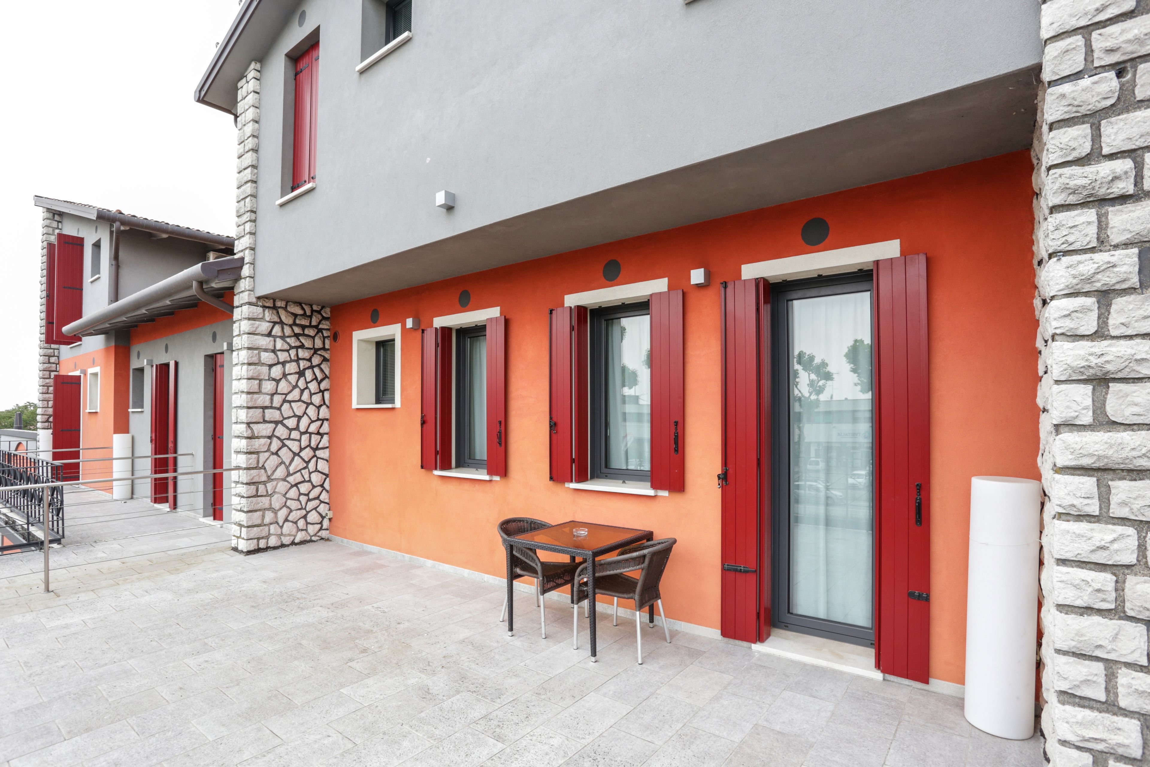 A modern building with a stone facade, red shutters, and a patio area with a table and chairs.
