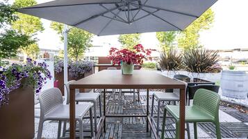 Een terras buiten met een tafel en stoelen, omringd door potplanten en een grote parasol.