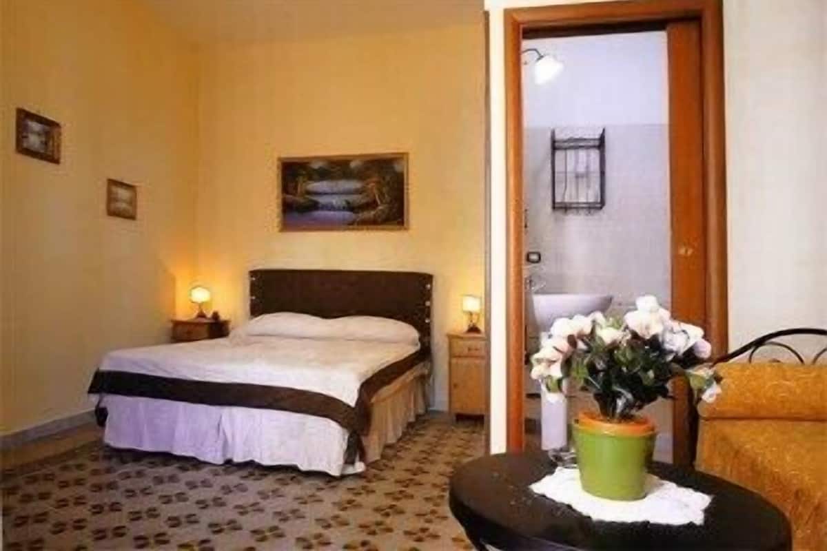 A hotel room with a bed, bedside lamps, a painting on the wall, a bathroom visible through an open door, and a small table with a vase of flowers.