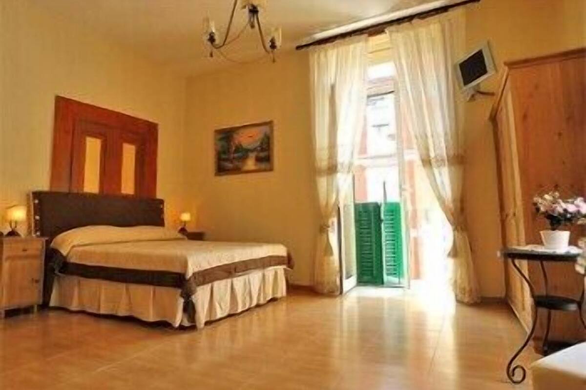 A bedroom with a bed, bedside tables, a painting, a television, and a view of the outside.