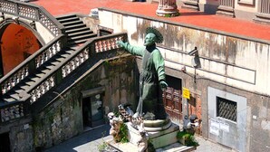 A statue of a historical figure on a pedestal with a staircase and a building in the background. - Domus Gemi (Naples)