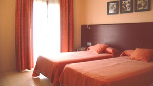 Une chambre d’hôtel avec deux lits, une tête de lit en bois et une fenêtre avec des rideaux. Trois tableaux encadrés sont accrochés au mur.