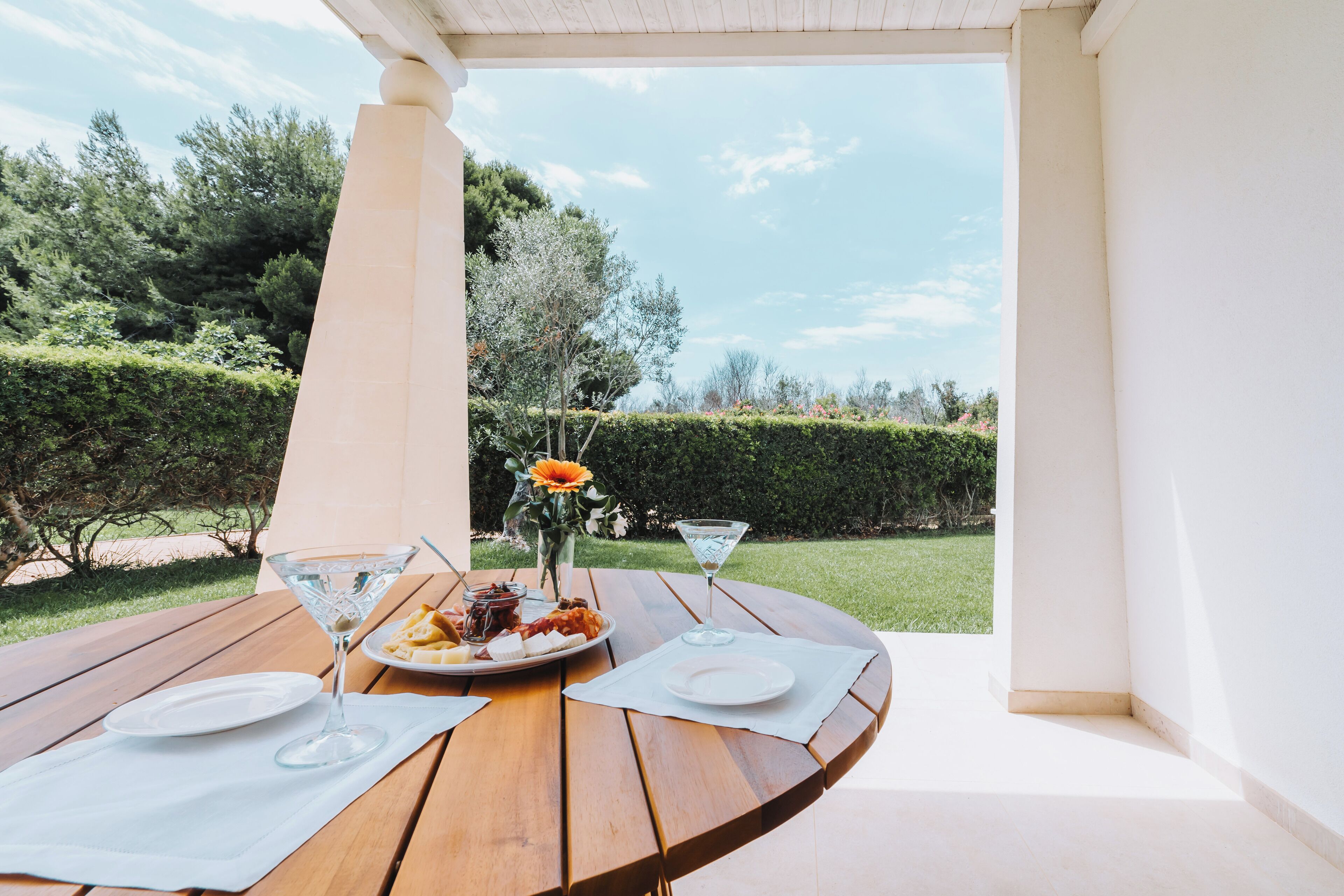 Une table en bois avec une nappe blanche, un verre à martini, une assiette de nourriture et un vase avec une fleur.