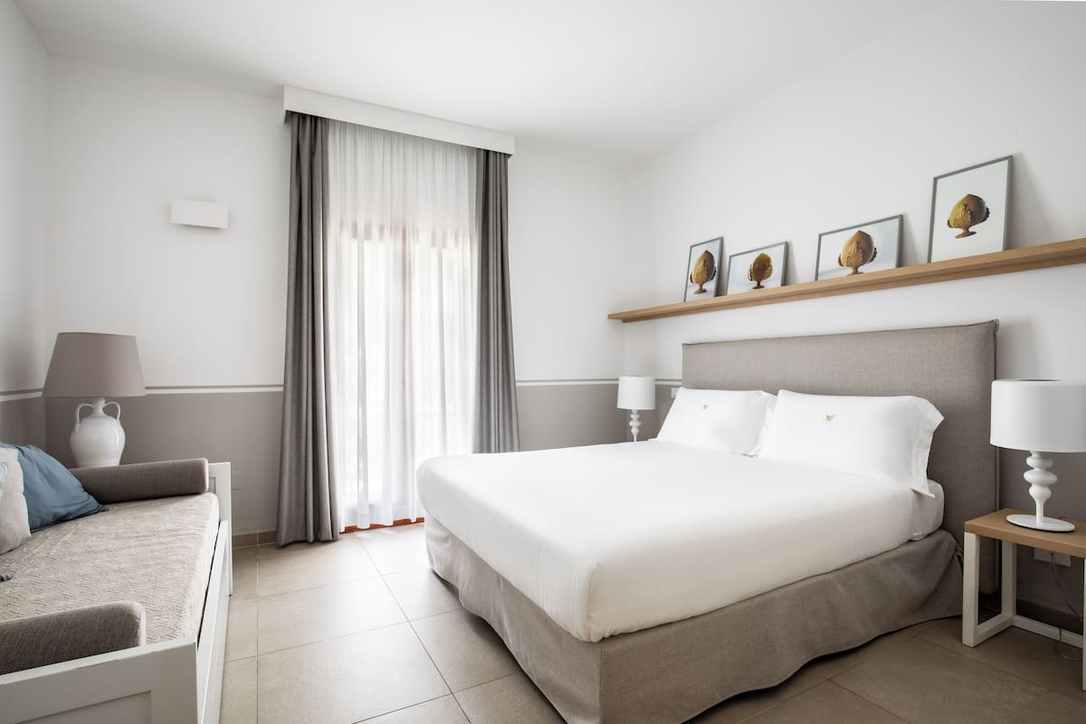 Una habitación de hotel moderna con una cama grande, una mesita de noche con lámpara, un pequeño sofá y una ventana con cortinas.
