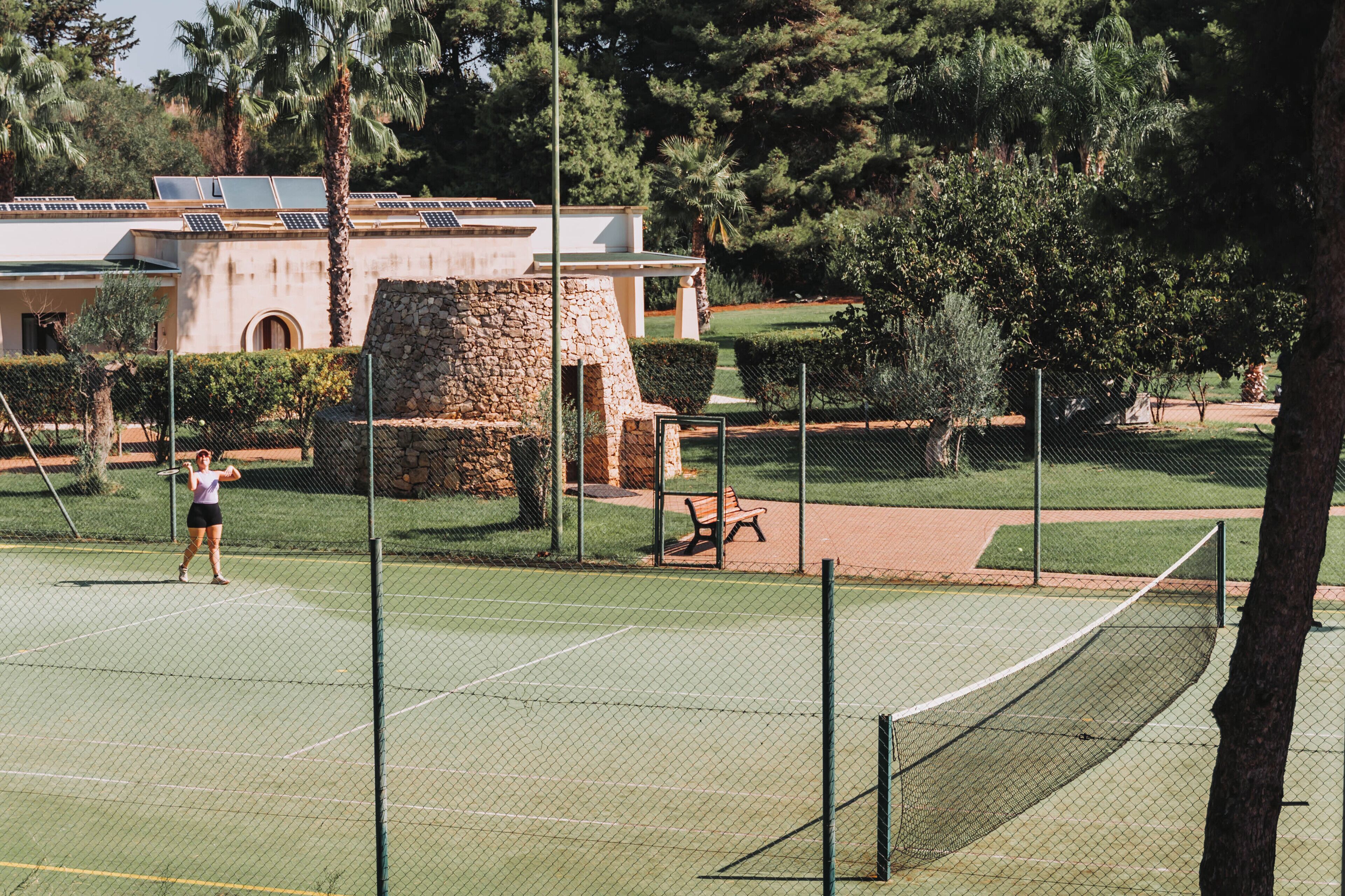 Un campo da tennis con una rete, circondato da una recinzione, e un edificio con facciata in pietra sullo sfondo.