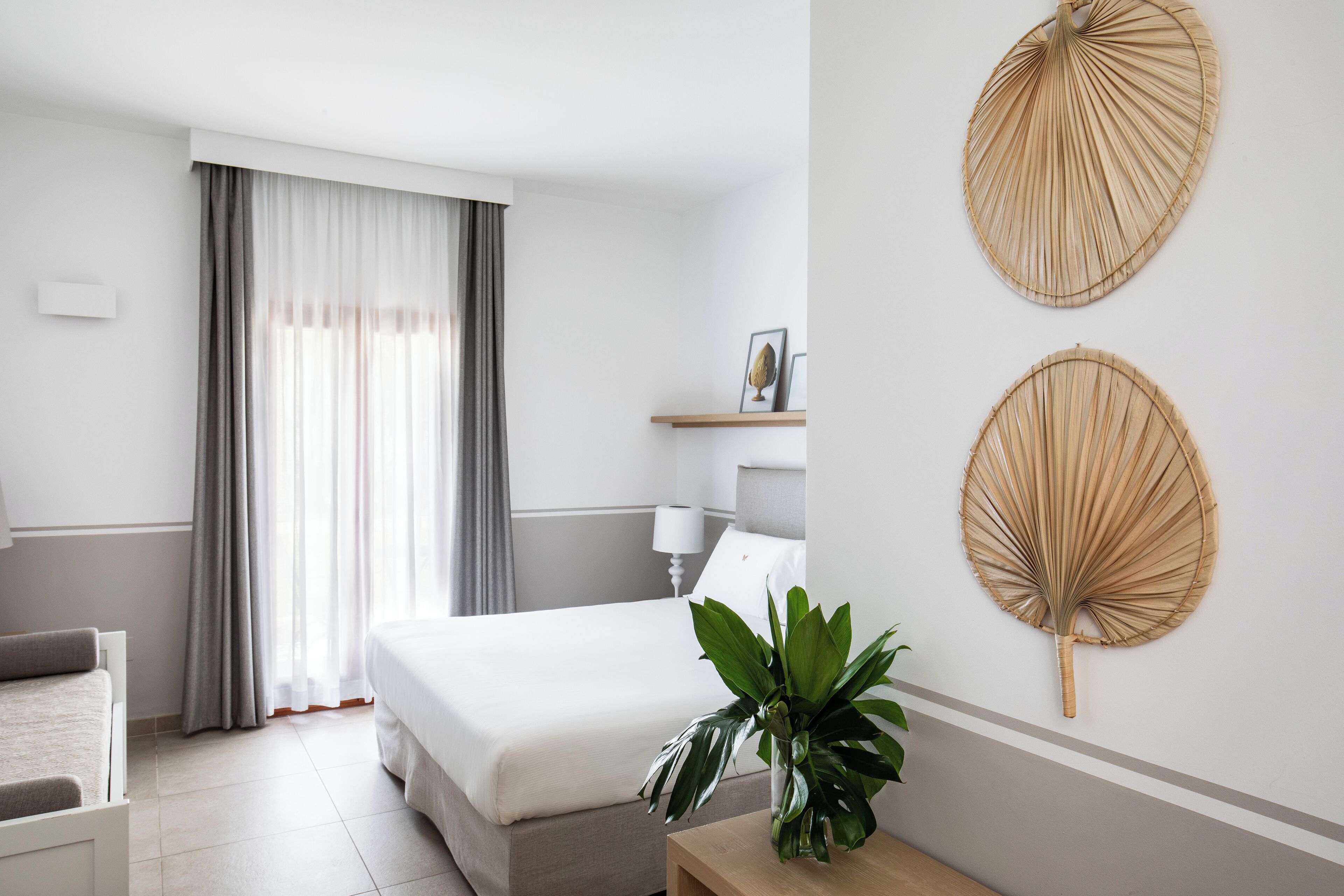 A hotel room with a bed, a bedside table with a plant, a bedside lamp, and two wall-mounted fans.