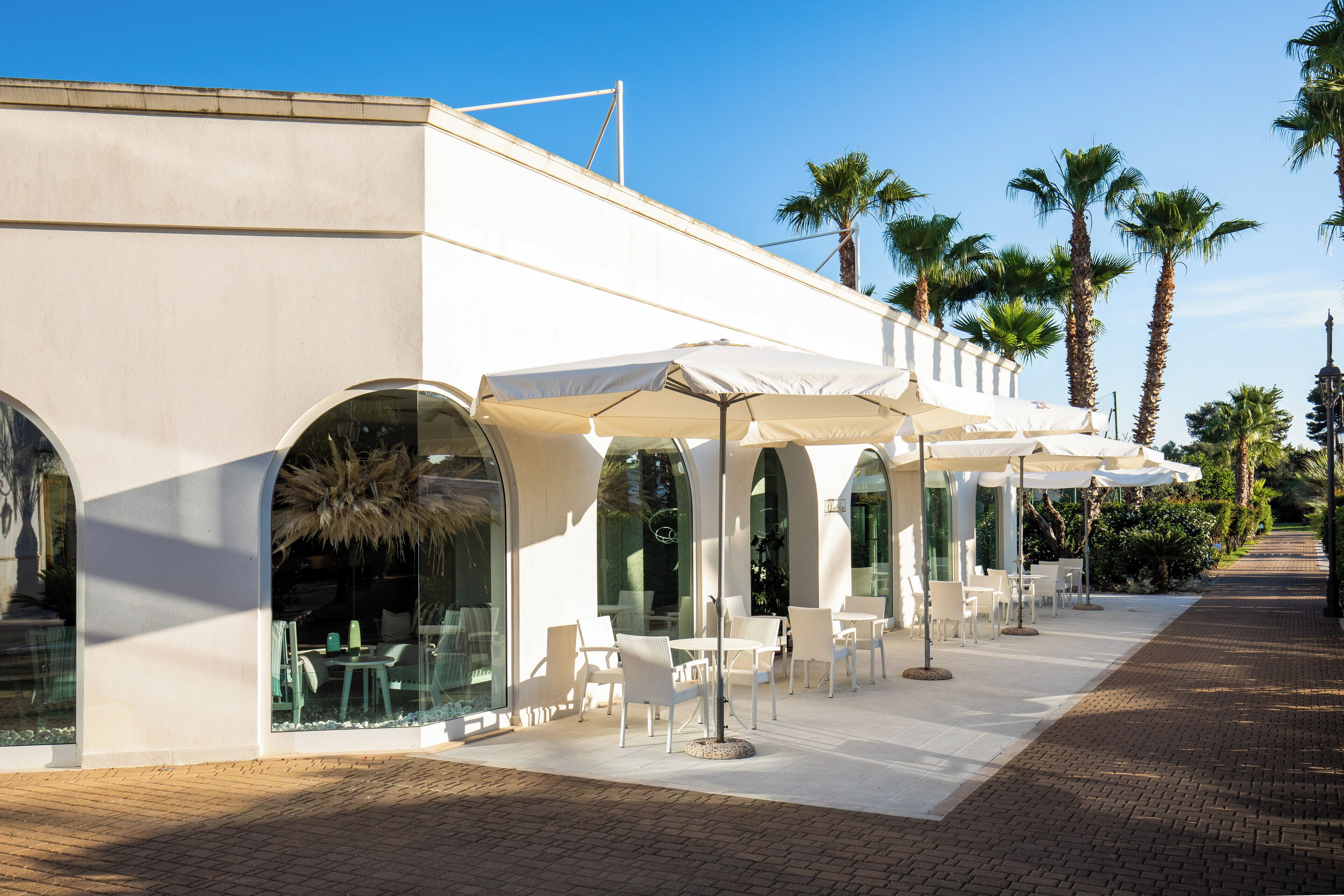 A modern outdoor cafe with white tables and chairs, covered by large umbrellas, and surrounded by palm trees.