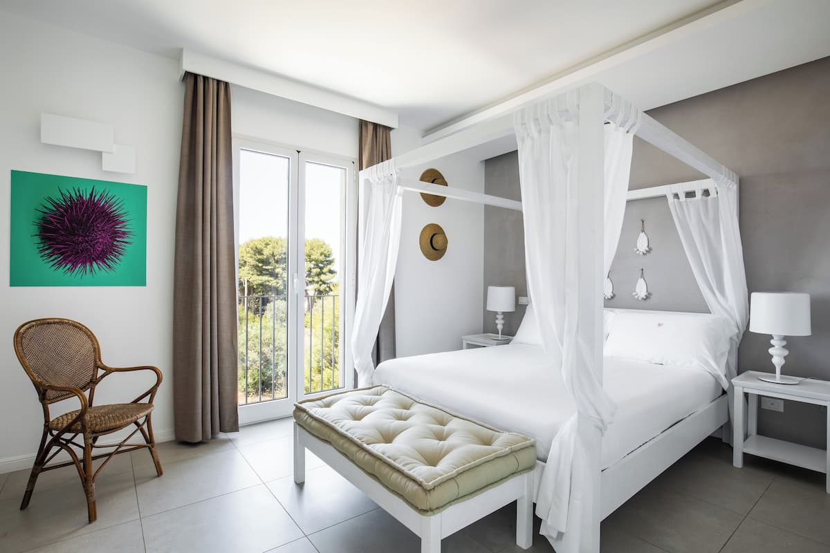 Un dormitorio con cama con dosel, una silla, una mesita de noche con lámpara y vistas al exterior.