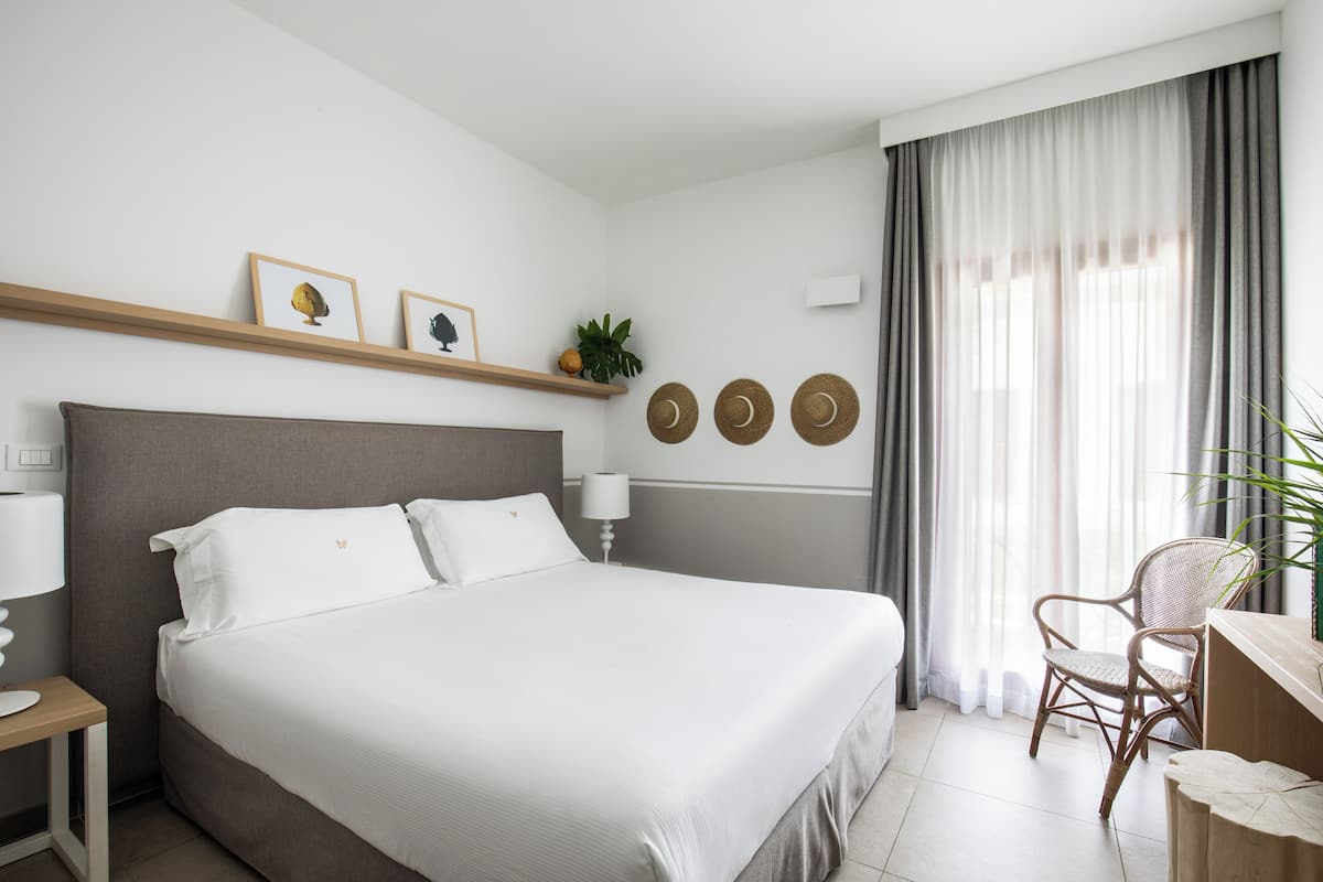 Una habitación de hotel ordenada con una cama grande, un escritorio con silla, una mesita de noche con lámpara y una ventana con cortinas.