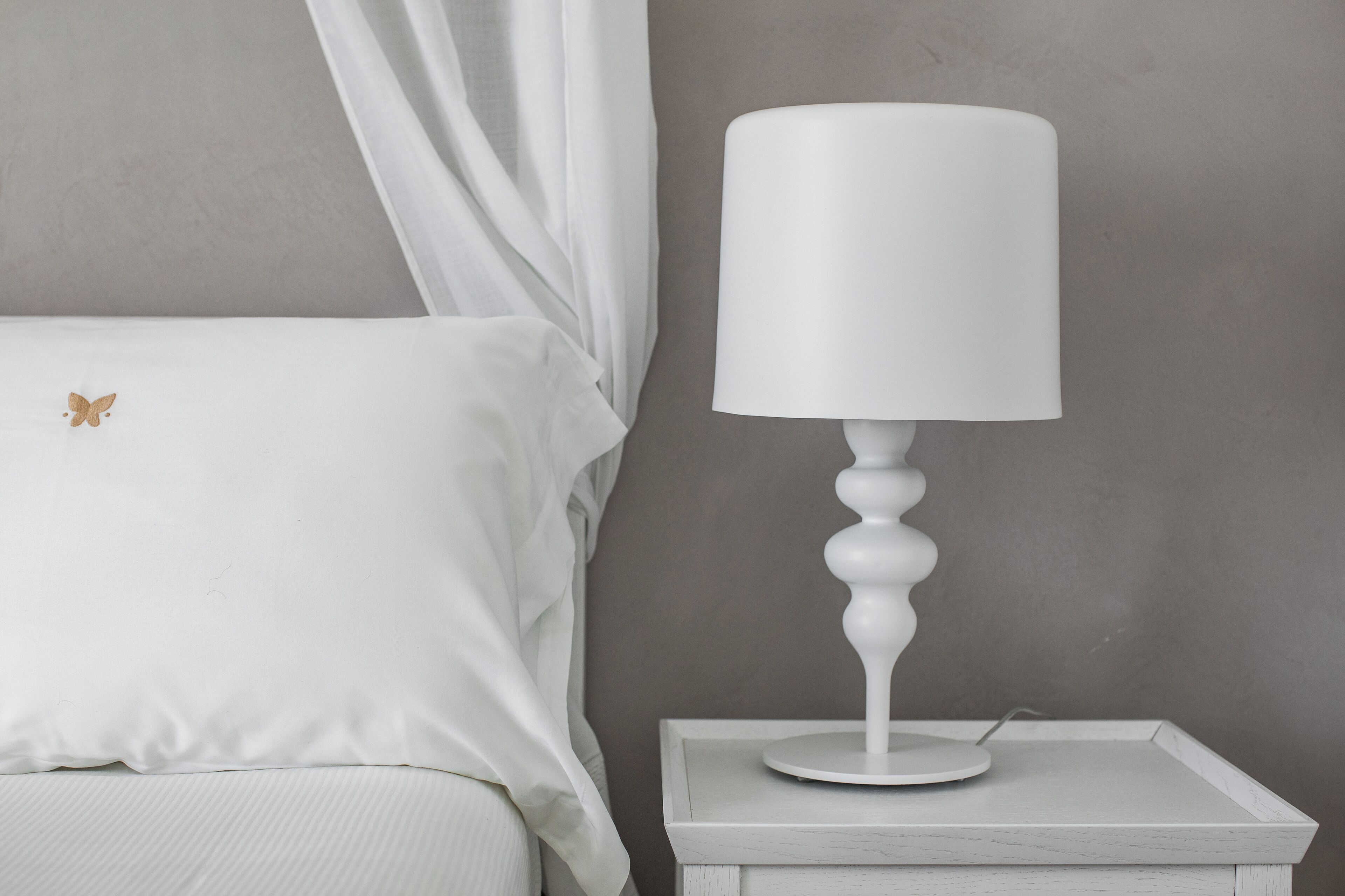 Une table de chevet blanche avec une lampe blanche, un lit blanc avec un coussin en forme de papillon et un mur gris.
