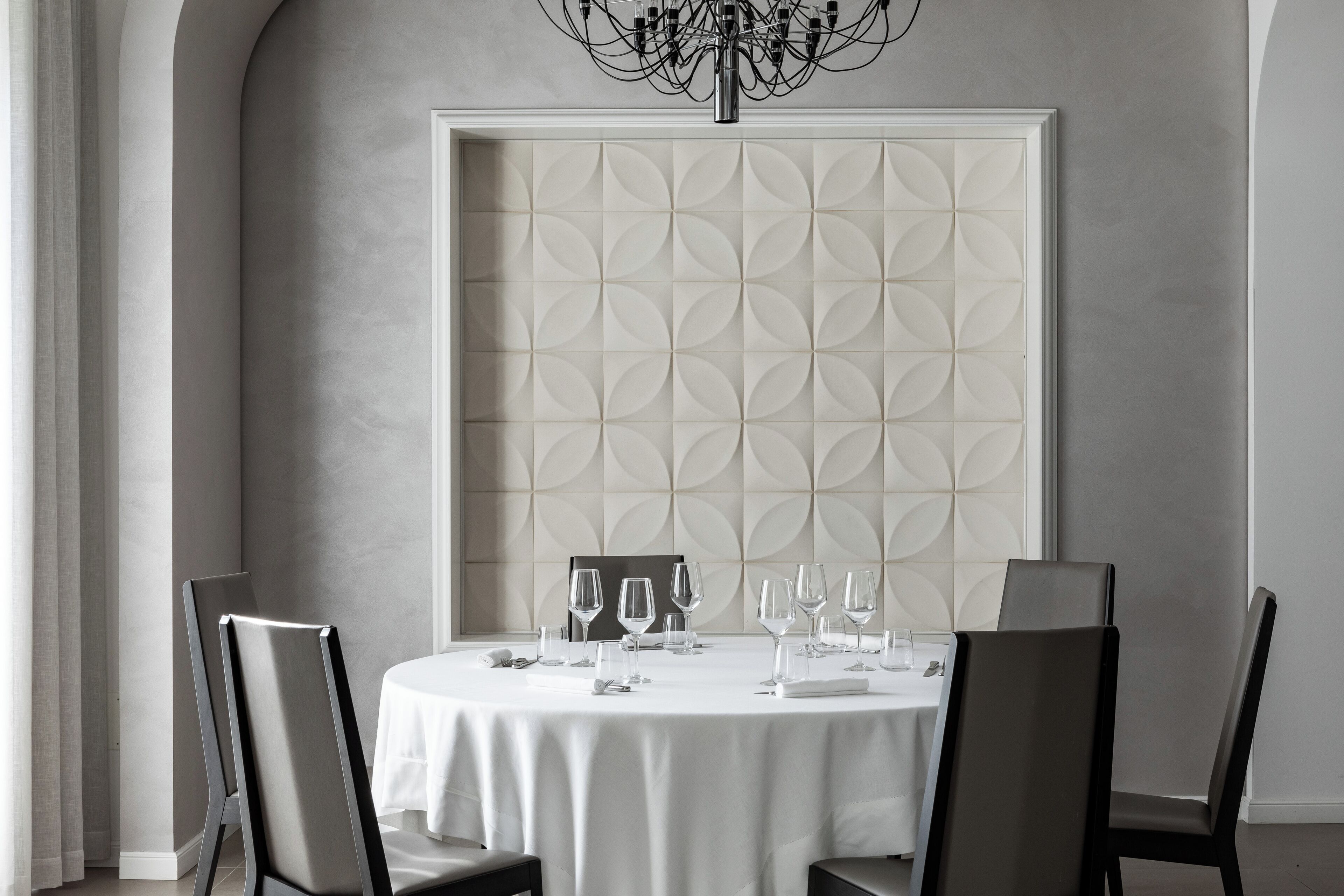 Una mesa redonda de comedor con mantel blanco, rodeada por cuatro sillas. Detrás de la mesa, una pared con diseño geométrico.