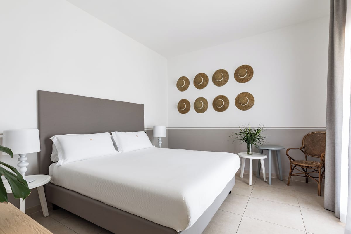Habitación de hotel ordenada con una cama grande, dos mesitas de noche con lámparas, una pequeña mesa redonda con una planta y una silla de mimbre.