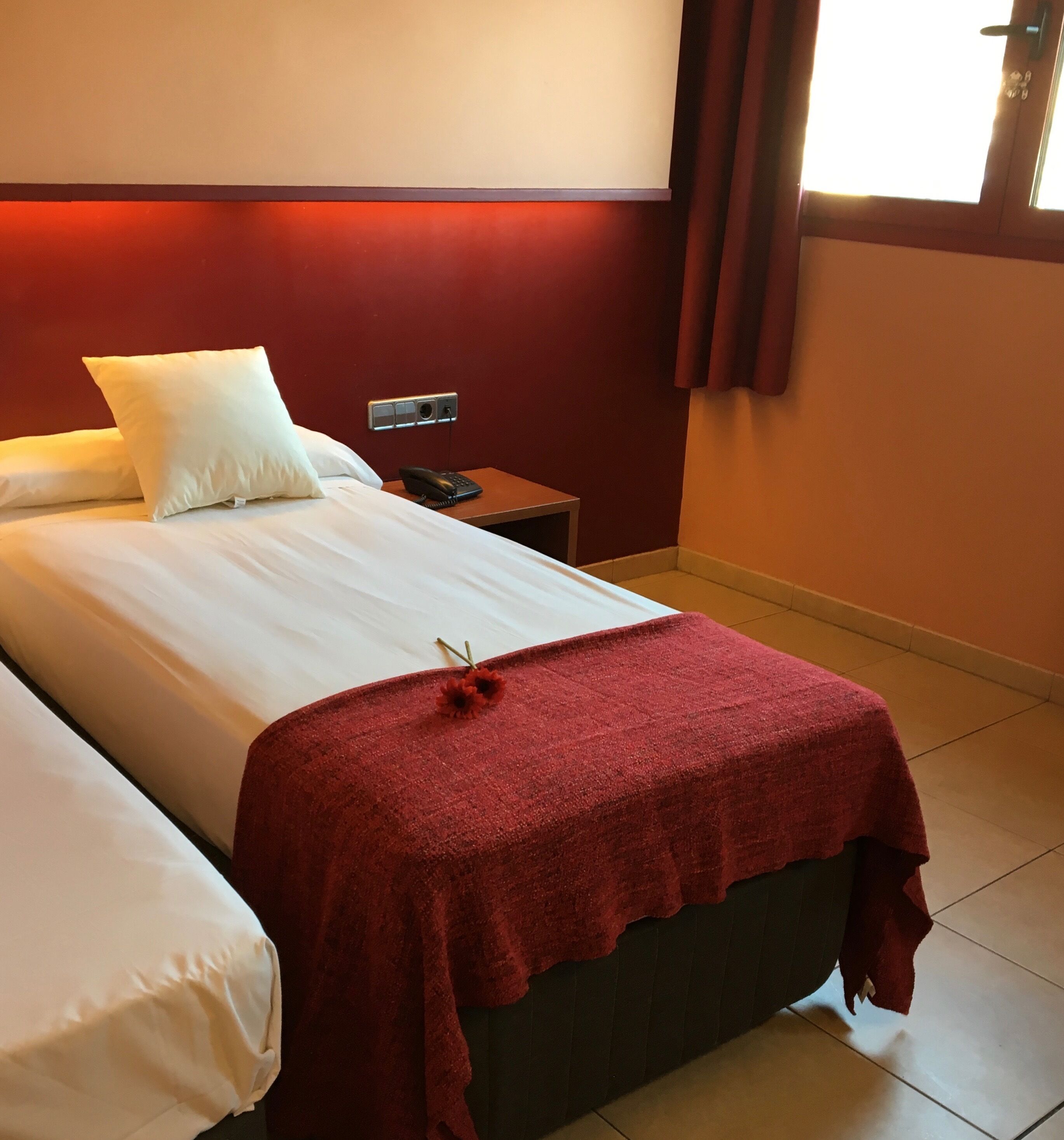 Una habitación de hotel con cama, mesita de noche, teléfono y ventana con cortinas.