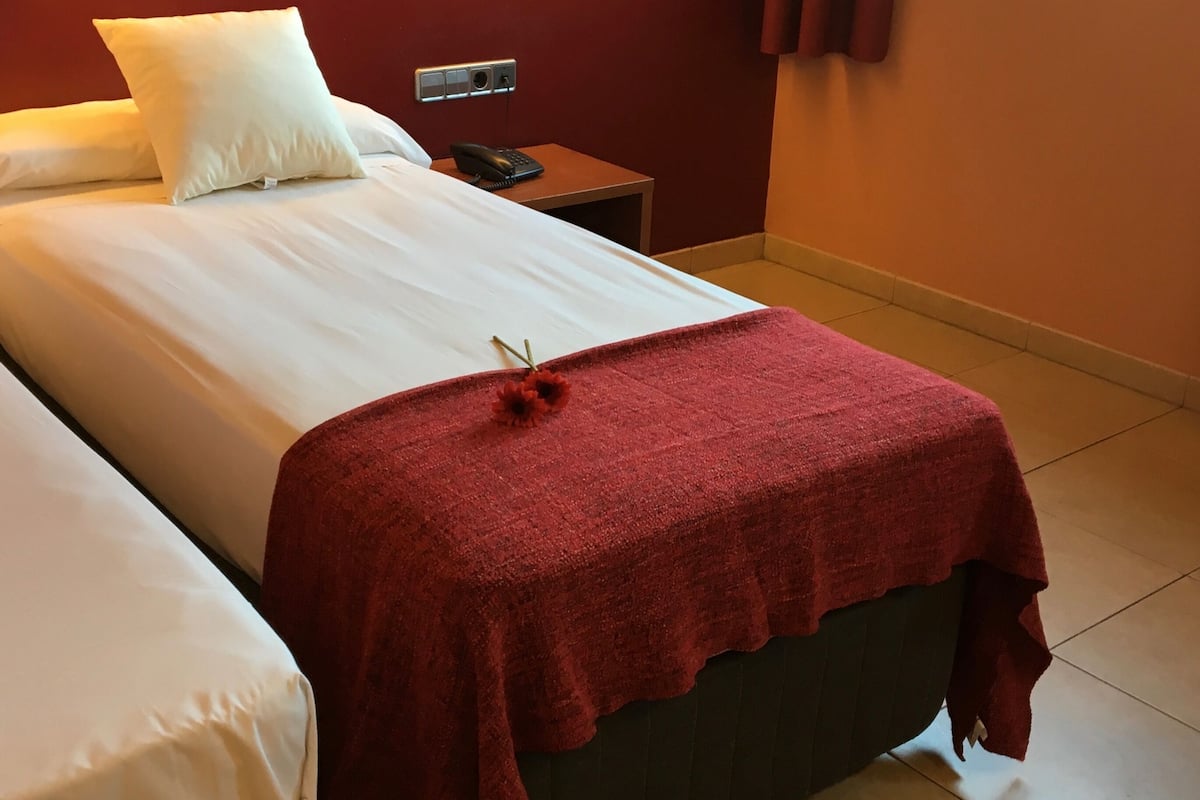 Una habitación de hotel con cama, mesita de noche, teléfono y ventana con cortinas.